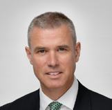Headshot of a smiling man with short grey hair, wearing a dark suit and a green patterned tie, against a plain light background.