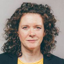 Headshot of a woman with curly brown hair, looking directly at the camera, against a light background.