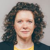 Headshot of a woman with curly brown hair, wearing a yellow top and dark blazer, against a plain light background.