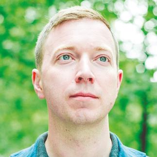 Headshot of a man with short blonde hair and blue eyes, looking slightly right, against a blurred green background.
