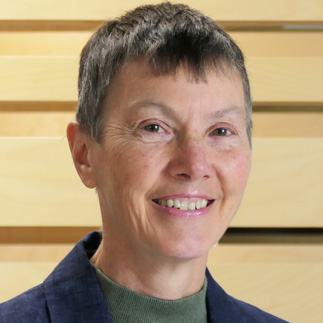 Headshot of a smiling woman with short grey hair, wearing a blue jacket, against a blurred wooden background.