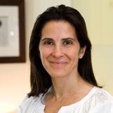 Professional headshot of a woman with long dark hair, smiling, wearing a white shirt, with a blurred indoor background.