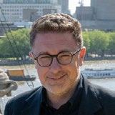 Professional headshot of a man with glasses and short gray hair, smiling, with a blurred cityscape and river in the background.