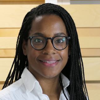 Headshot of a smiling woman with long braided hair and glasses, wearing a white shirt, against a blurred wooden background.