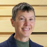 Headshot of an older woman with short grey hair, wearing a green top and dark blue jacket, smiling.