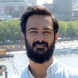 Professional headshot of a man with a beard and dark hair, smiling, with a blurred cityscape and river in the background.