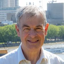 Headshot of an older man with gray hair, smiling, against a blurry cityscape.