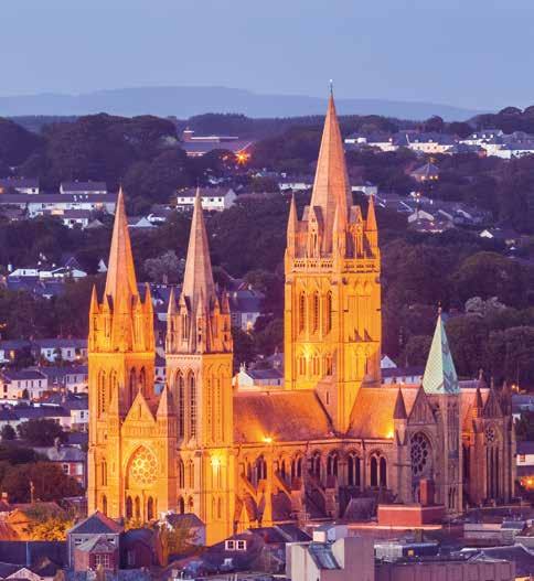 St. Fin Barre's Cathedral in Cork, Ireland, illuminated at dusk with city lights in the background.