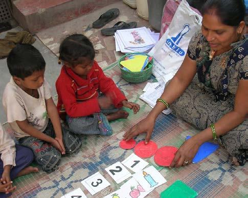 A woman and two children sit on a patterned mat, engaging in a learning activity with number cards and red circular cutouts.