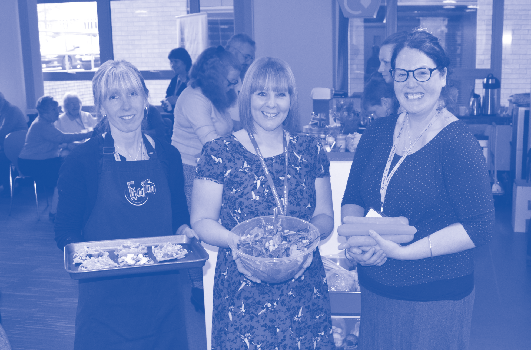 Three smiling women hold food items, including a salad bowl and a tray of baked goods, in a communal setting. Grow, Share, Cook Cities of Service, Plymouth City Council.