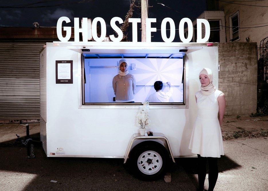 A white food truck with "GHOSTFOOD" sign, showing two people dressed in white, one inside and one outside, against a dark street background.