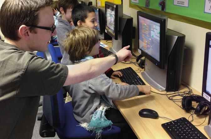 An adult male assists two young boys at computers, pointing at a screen in a classroom setting.