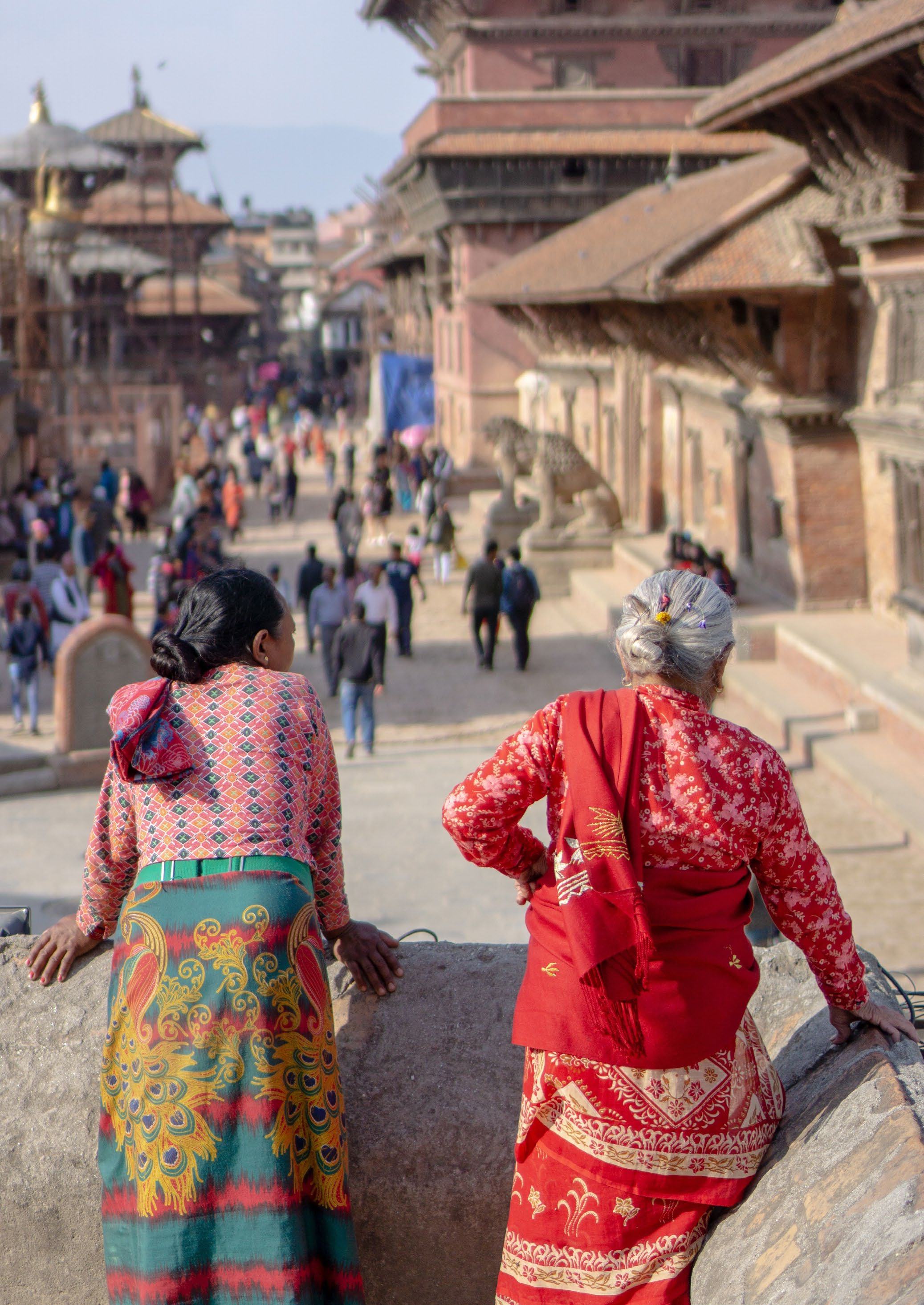 Two women in traditional attire overlook a bustling street lined with historic multi-tiered buildings in an old city.