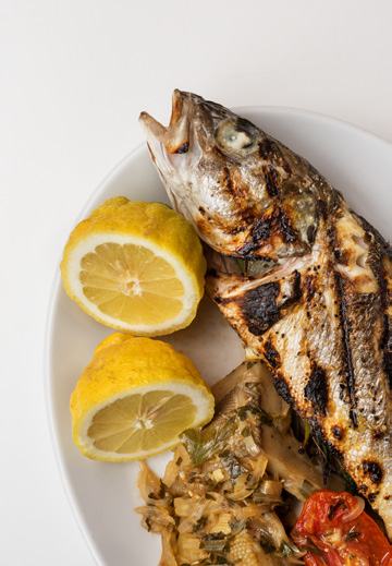 A white plate holds a perfectly grilled whole fish, two lemon halves, and a serving of sautéed vegetables.
