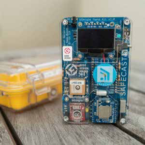 Product photo of a blue bGeigie Nano Kit v1.0 circuit board with "SAFECAST" branding, next to a blurred yellow protective case.