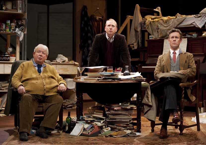 Three men on a stage set: one seated left, one standing behind a cluttered desk, and one seated right with a briefcase.