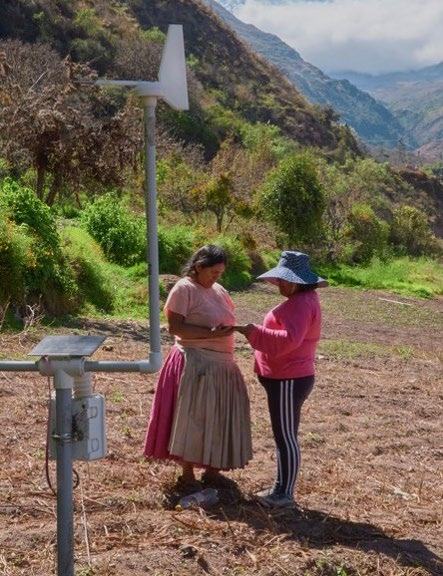 Two women observing a scientific sensor station in a field with mountains in the background, possibly collecting environmental data.