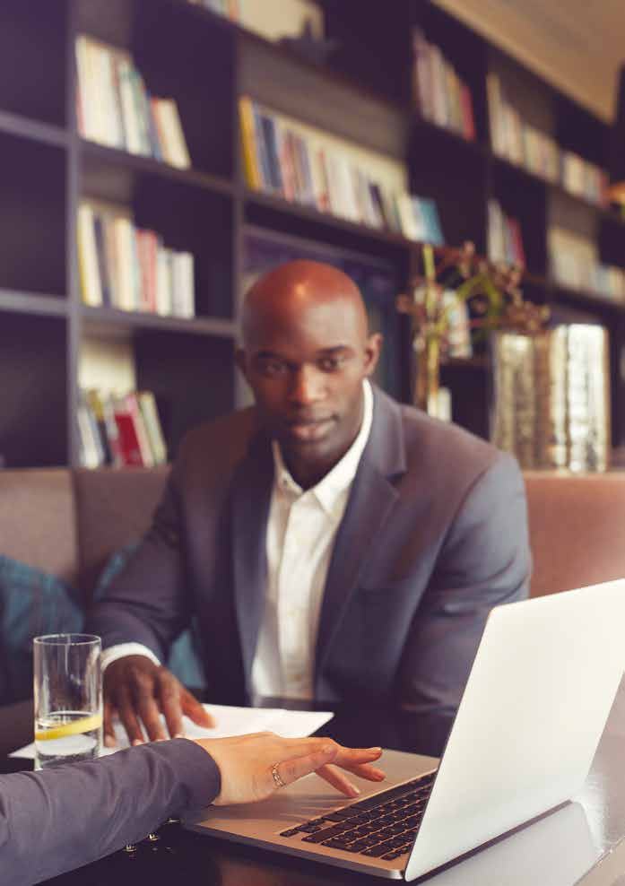 A businessman in a suit at a table with a laptop, engaging with another person's hand gesturing towards the screen in a modern office.