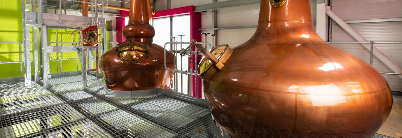 Two large, polished copper pot stills stand on a metal gridded platform inside a modern distillery.