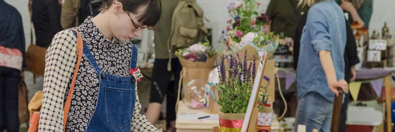 A person in glasses and overalls looking at items on a stall, with potted lavender and flowers in the background.