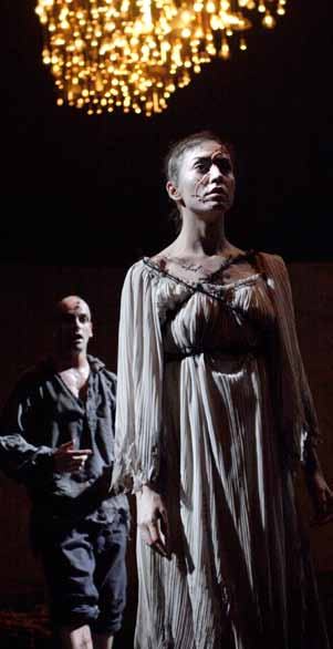 A woman with pale makeup and facial marks looks up on a dark stage, with a man blurred behind her and a chandelier above.
