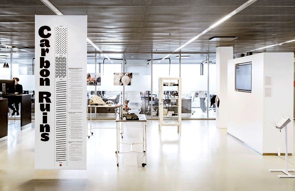 Exhibition space with a tall banner titled "Carbon Ruins" featuring text and small graphics, alongside display cases and screens.
