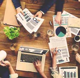 Overhead view of a business meeting with multiple hands interacting with documents, charts, a laptop, and coffee cups on a wooden table.