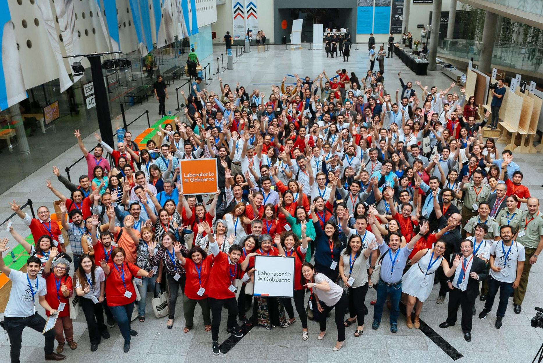 A large group of people smiling and waving, holding "Laboratorio de Gobierno" and "Experimenta" banners, indicating a successful event or gathering.