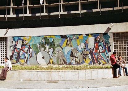 Outdoor mosaic art depicting abstract figures and industrial themes on a building wall, with people sitting on a planter in front.