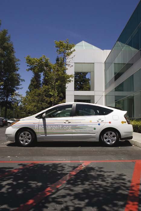 A white hybrid car, decorated with colorful stripes and "Rechargeit.org" branding, parked in front of a modern office building.