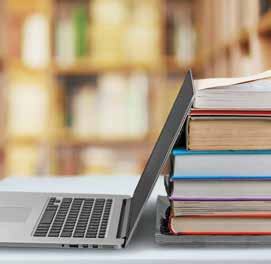 An open laptop stands next to a stack of textbooks on a desk, with a blurred library background, symbolizing education or digital learning.