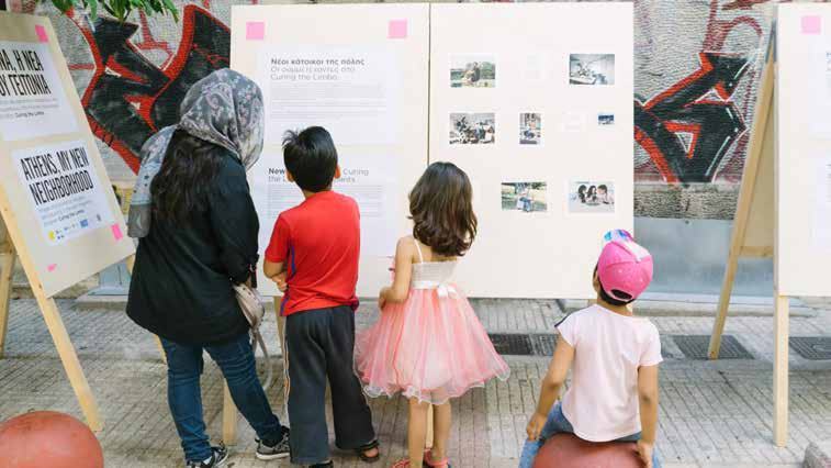 People observing an outdoor exhibit with informational boards and photographs, titled 'Athens, My New Neighborhood'.