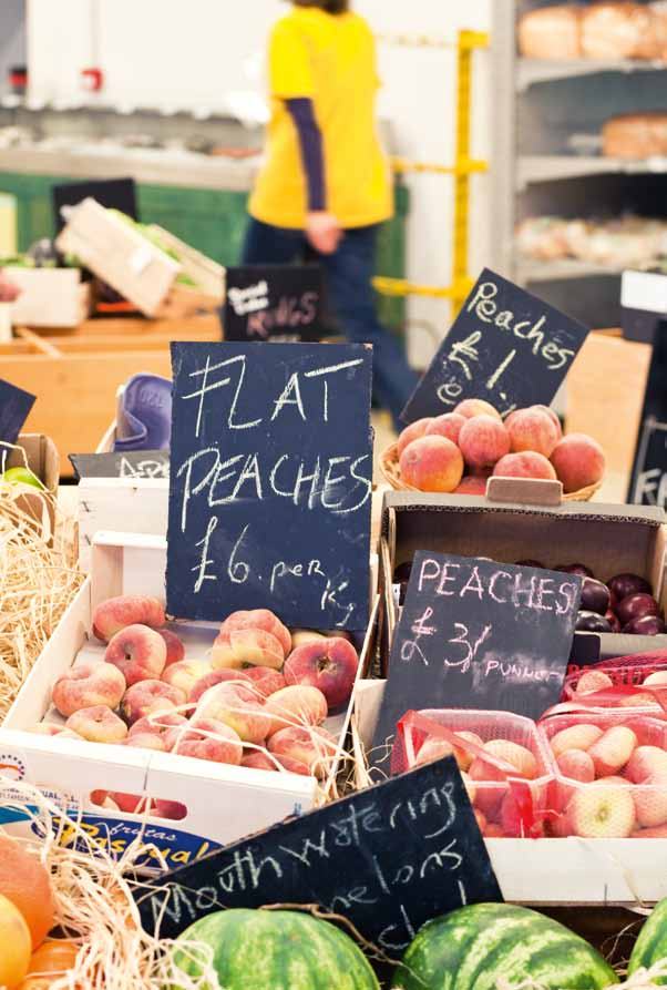 A market stall with peaches, watermelons, and chalk signs showing prices like "Flat Peaches £6 per Kg" and "Peaches £3 / Punnet".