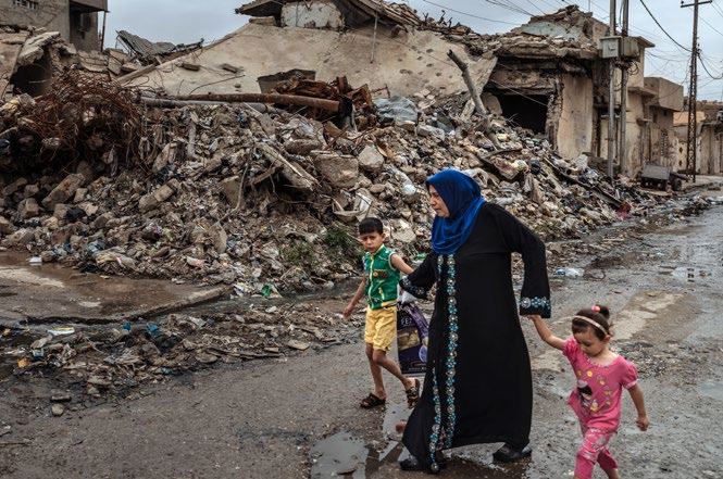 A woman in a hijab holds hands with two children, walking past a large pile of building rubble and damaged structures.