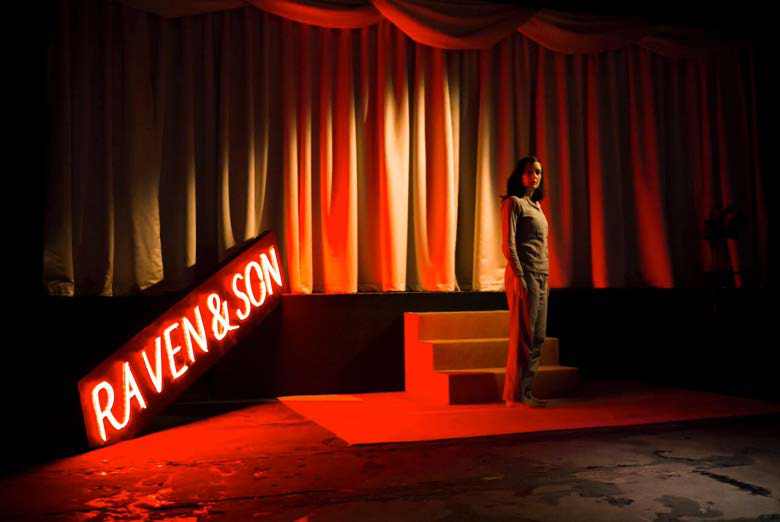 A person standing on a stage with a red neon sign reading "RAVEN & SON" and dramatic red lighting.