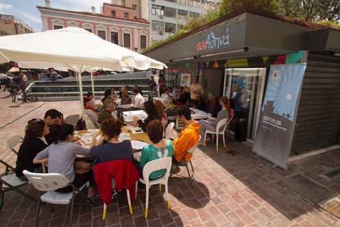 People gathered at outdoor tables near a synAthina kiosk, participating in an event or meeting.