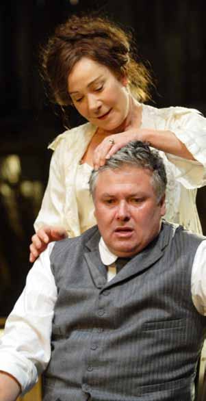 A woman stands behind a man, gently placing her hands on his head as he looks forward with a distressed expression.