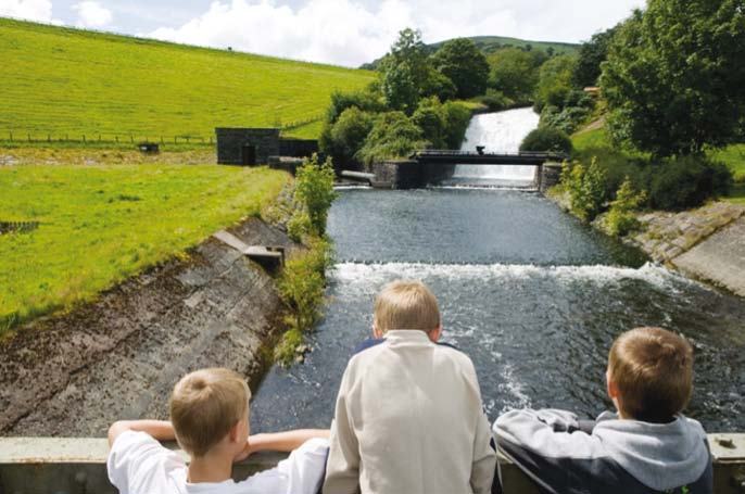 Three boys looking over a railing at a river flowing over a small dam or weir, surrounded by lush green hills.