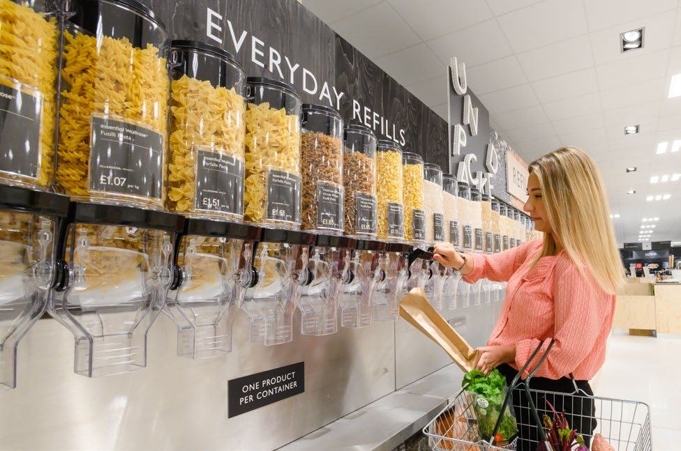 A woman refills pasta from a bulk dispenser in a supermarket aisle marked "EVERYDAY REFILLS," showing products with prices and packaging.