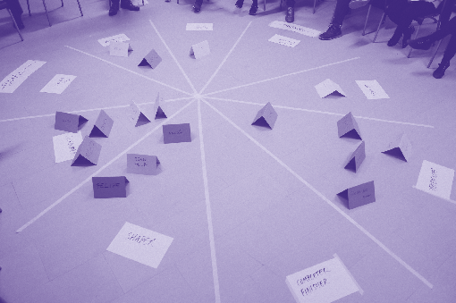 Overhead view of a workshop activity with a circular diagram on the floor divided into wedges, containing folded paper labels.