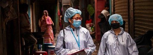 Two people in medical gowns, masks, and hairnets walk down a narrow, crowded street, appearing to be conducting fieldwork.