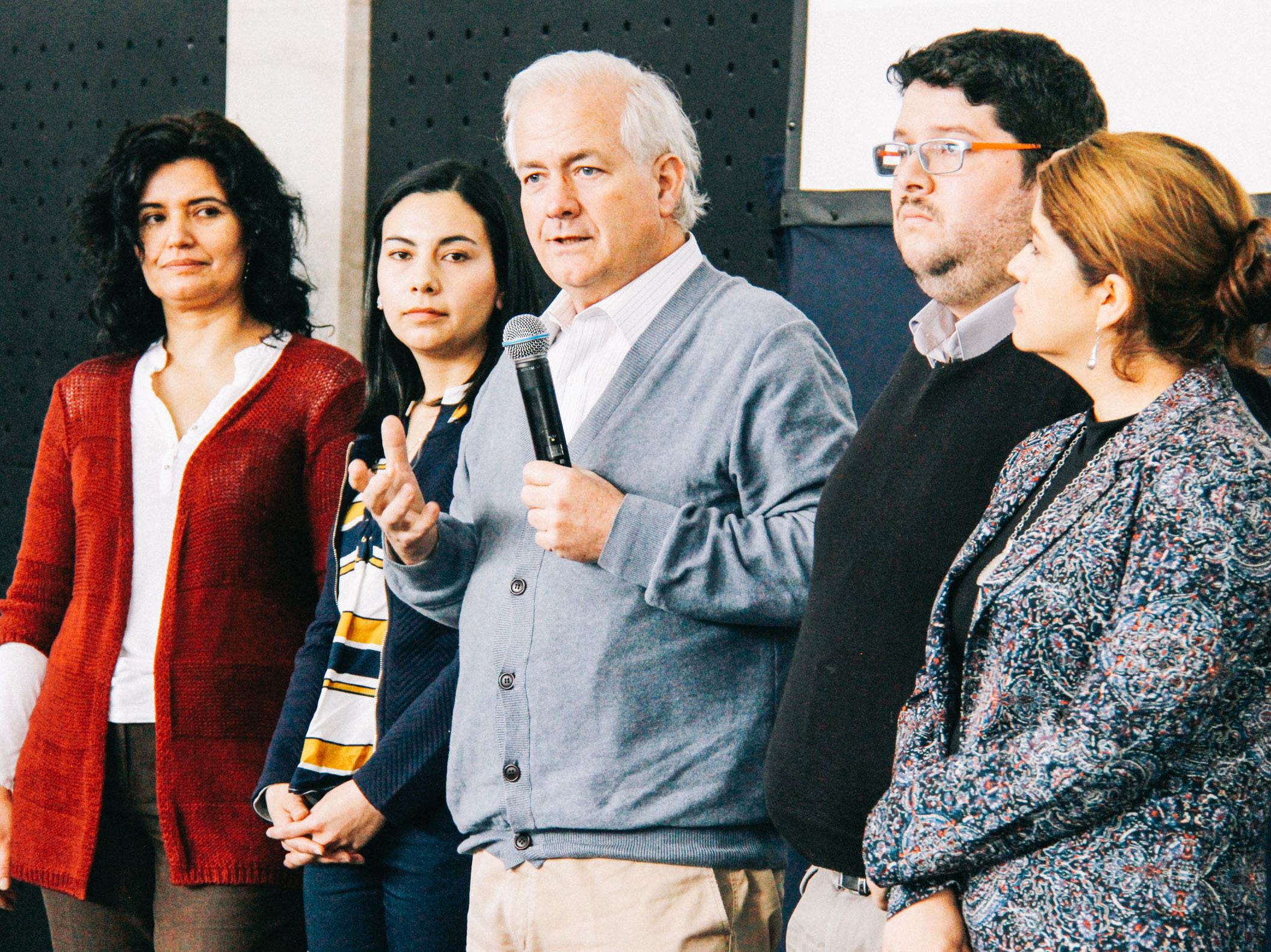 A man holding a microphone speaks to an audience, flanked by four other individuals, suggesting a presentation or panel discussion.