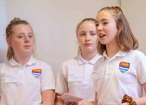 Three young women in white polo shirts with a crest logo, one speaking to an unseen audience.