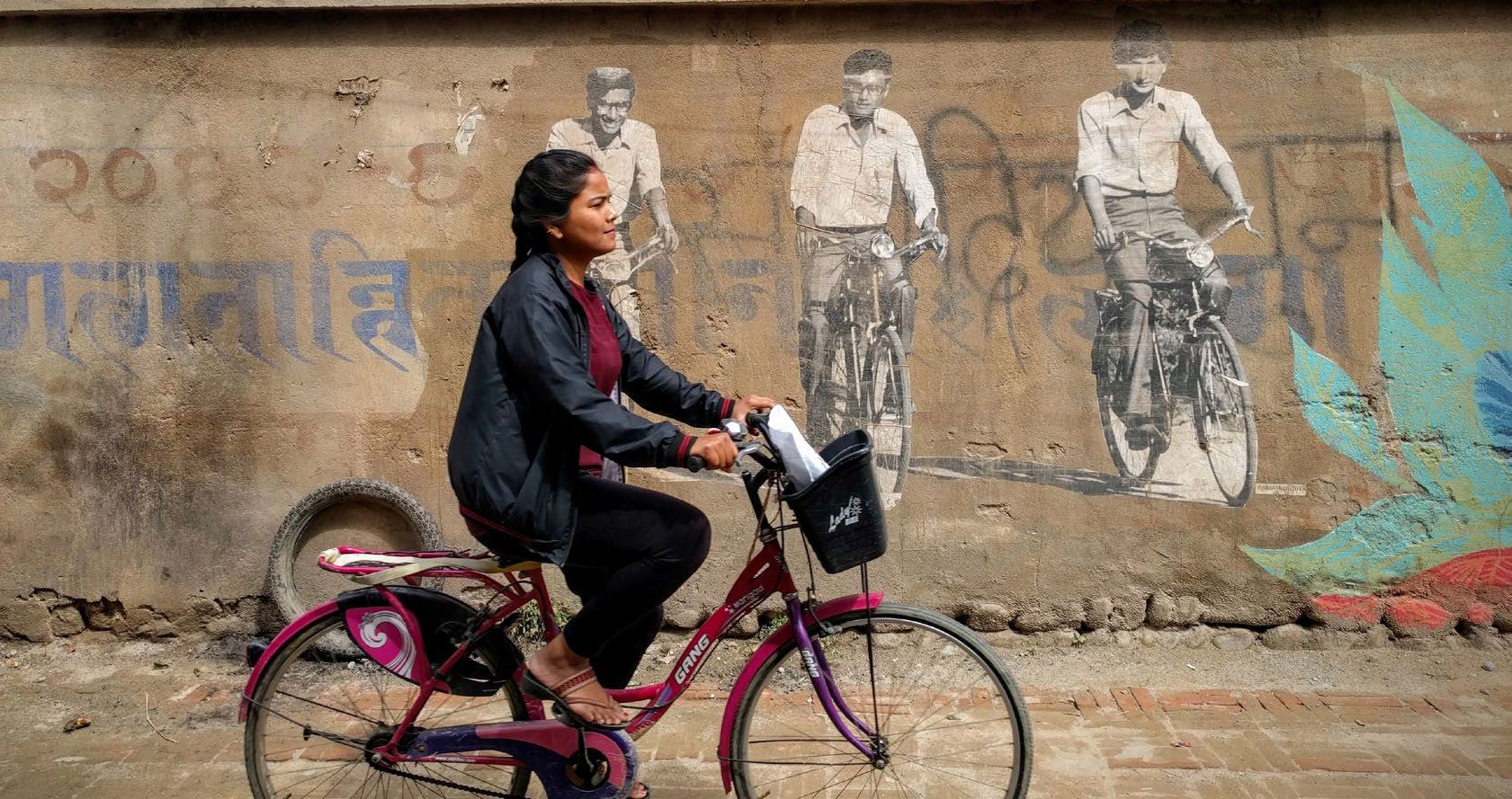 A woman rides a pink bicycle down a street next to a wall with murals of people on bicycles.