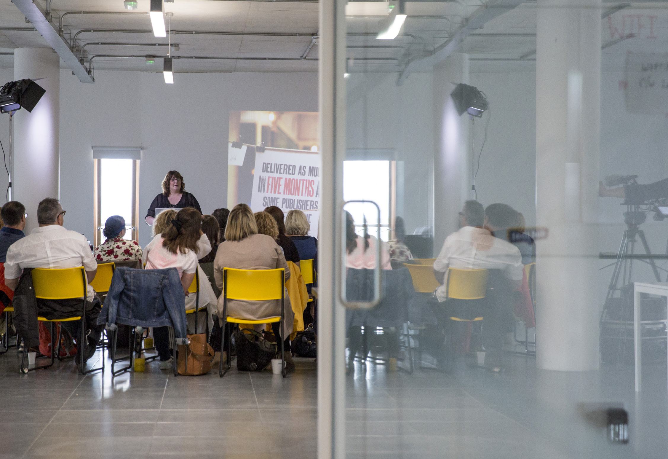A woman presents to a group in a brightly lit room, with text visible on a projector screen, partially seen through a glass partition.