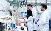 Three scientists in lab coats working with complex experimental equipment and monitors in a laboratory setting.