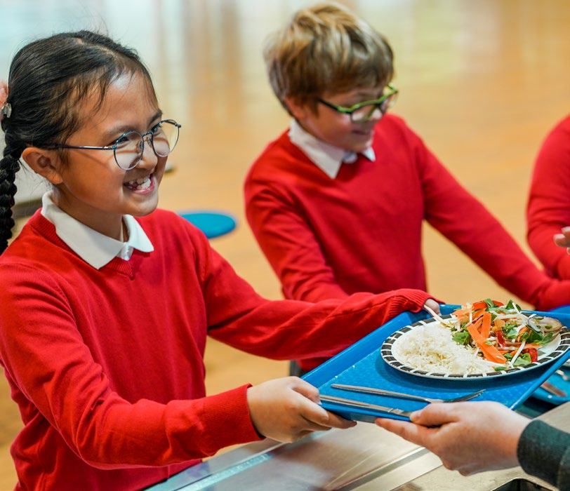 A smiling schoolgirl in red uniform receives a healthy meal tray in a cafeteria.