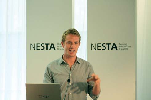 Man speaking at a presentation, standing at a podium with a laptop, against a backdrop with "NESTA Making Innovation Flourish" logos.