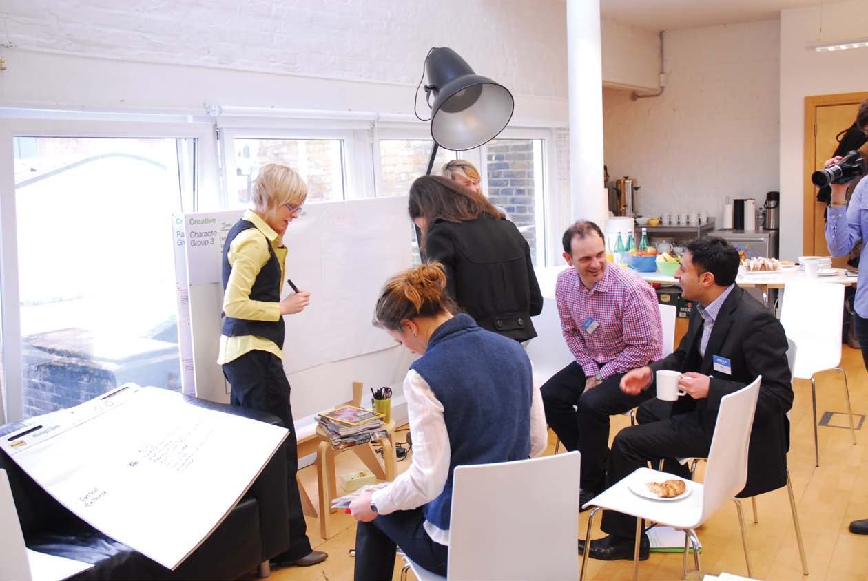A group of people engaged in a workshop, with one woman writing on a whiteboard and others observing or taking notes in a bright room.