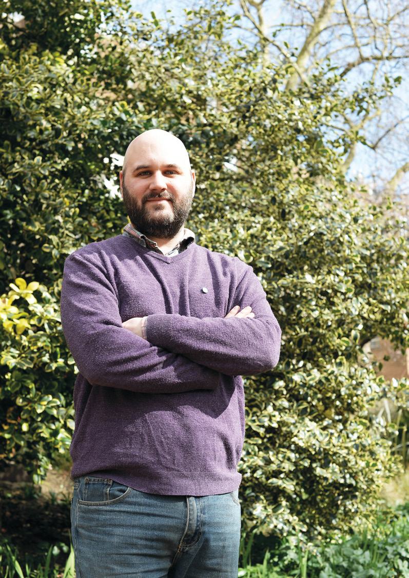 A man with a beard and a purple sweater stands outdoors in front of green foliage, arms crossed.